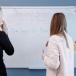 Two students studying English grammar at a whiteboard in a classroom setting.